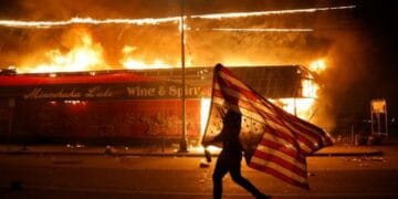 A protester carries a U.S. flag upside, a sign of distress, next to a burning building Thursday, May 28, 2020, in Minneapolis. Protests over the death of George Floyd, a black man who died in police custody Monday, broke out in Minneapolis for a third straight night. (AP Photo/Julio Cortez)