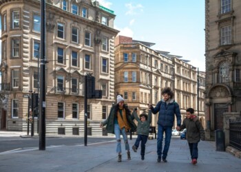 A multiracial family of four, a mother, father and their two little boys on a staycation in the city of Newcastle on a sunny winters day. They are wearing casual, winter clothing and accessories. They are walking down a city street, holding hands and swinging one of the boys in the air.