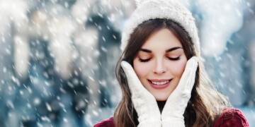 Outdoor close up photo of young beautiful happy smiling girl walking on street. Model closed her eyes and touching face, wearing stylish white knitted winter hat and gloves. Copy, empty space for text