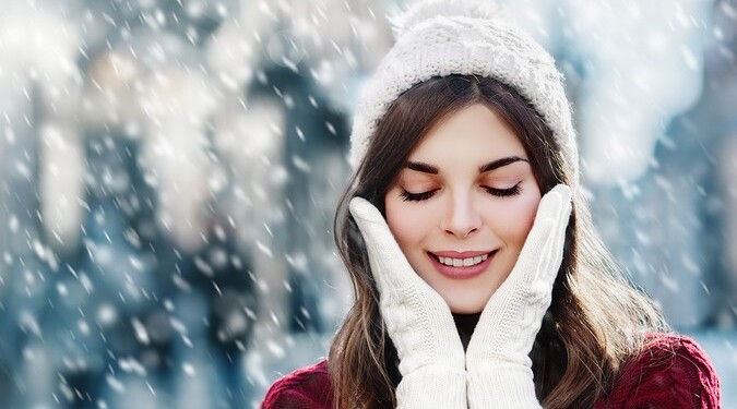 Outdoor close up photo of young beautiful happy smiling girl walking on street. Model closed her eyes and touching face, wearing stylish white knitted winter hat and gloves. Copy, empty space for text