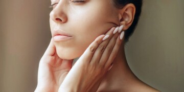 Cropped shot of young caucasian woman touching skin with hands massaging her face isolated on a dark brown background. Rejuvenation, facelift, facefitness. Cosmetology and beauty concept