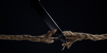 Horizontal shot of stretched cord is cut with a knife on a dark background. The rope is about to break. The concept of the danger of rupture.