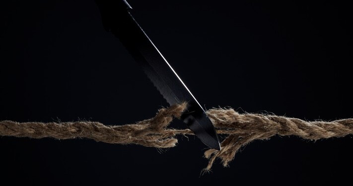 Horizontal shot of stretched cord is cut with a knife on a dark background. The rope is about to break. The concept of the danger of rupture.