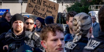 Police officers stand next to demonstrators with a sign "no Friedrich, the AfD is no alternative" during a protest against an AfD election campaign in Neu Isenburg near Frankfurt, Germany, Saturday, Feb. 1, 2025. (AP Photo/Michael Probst)